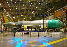 Hall d'assemblage d'avions, avec un appareil biréacteur non peint, un podium de présentation et des chaises disposés pour le public.