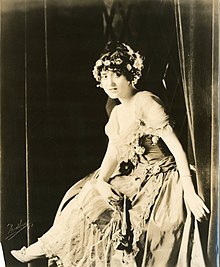 A young white woman, seated, wearing a flower crown and a gown.