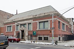 A square two-story brick building stands before a snowy sidewalk against a gray sky.