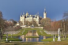 Frontale Farbfotografie eines Schlosses im Renaissance-Stil mit mehreren kleinen Türmen und einer goldenen Kuppel. Im Vordergrund sind an einer angelegten, langen Wasserfläche Uferwiesen und aufgereihte, kleine Bäume. Im Wasser ist eine runde Rasenfläche. An den Ufern stehen mehrere Skulpturen auf Sockeln.