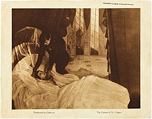 Un lobby card avec une photographie au centre, et de petites légendes en dessous. L'image représente un homme vêtu de noir se pencher vers le lit blanc d'une jeune femme endormie.