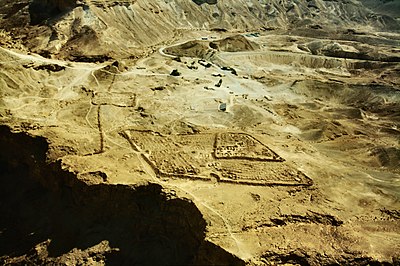 Der Blick geht über eine Wüstenlandschaft, aus der sich antike Strukturen abzeichnen. Im Zentrum sieht man das Parallelogramm eines römischen Lagers, dahinter verläuft der Belagerungswall, in den ein kleines Lager mit etwa quadratischem Grundriss einbezogen ist