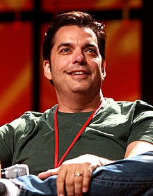 Photo d'un homme souriant, en position assise, portant un t-shirt vert et tenant un micro