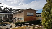 Andrews building is two-storeys high, and consists of a long grey stucco building and a shorter brown brick building which comes off of it at a right angle.