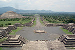 A large stone pyramid with smoothed surfaces and stairs leading to the top. Smaller pyramids of about 4 levels are surrounding a large square and line a wide road.