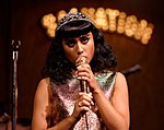 Photographie montrant une femme aux cheveux noirs, Natalia Kills, portant une robe argentée et empoignant un microphone doré.