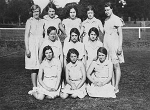 A black and white photo of a netball team. All the girls in the picture are school aged, wearing white and have their netball skirts on. The skirts are knee length and the girls are standing in three rows, with the front row sitting and the middle row kneeling.