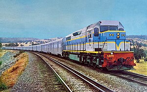 In a rural scene, a two-tone blue diesel locomotive with yellow striping and red headstock at the head of the Indian Pacific train comprising stainless steel passenger cars