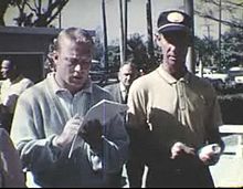 "Two young men, one blond and one and one in a cap standing next to each other while the blond man signs an item for the other man."