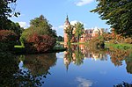 A red, ornate neo-gothic castle in a park-like location, the main tower of the castle is located to the left and topped with an ornate round dome and spire