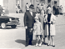 Photo en noir et blanc avec quatre personnes au premier plan, un homme, deux femmes et une jeune fille. L'homme sur la gauche porte un costume et un chapeau.