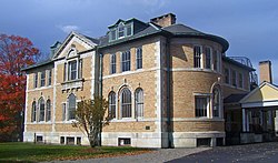 A golden brick building with a rounded wing on the right and pointed section in the middle of its roofline. It has lighter trim on the building, rounded windows on the first floor, and green trim on its black roof.