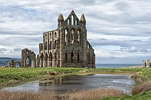 Photo d'une grande église en ruines, dépourvue de toit, qui surplombe un étang au premier plan et la mer à l'horizon