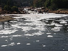 Ao fundo, a água corre por entre as pedras e, mais a frente, corre calma e lentamente. Sobre a água, uma camada de espuma branca.