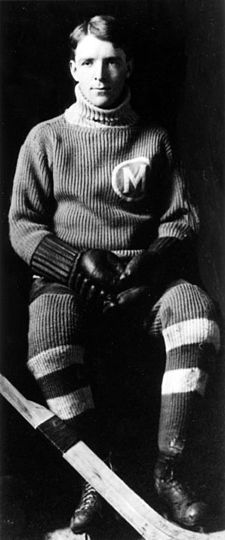 A young man in full hockey gear sitting. He has short hair parted on his right side and is wearing a dark uniform with a light coloured "M" logo in a circle on his left breast