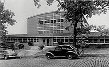 A brick building with many windows, with a brick street in front of it. There is a large tree on the right side of the image, in front of the building. There are two cars parked next to the building, one in the center and one on the left edge of the image. The building's entrance has the word "Research" above it.