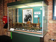 Una vitrina de vidrio que contiene un modelo grande de latón de una locomotora de vapor. En la parte posterior de la caja hay una fotografía en blanco y negro de un hombre.