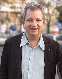 Homme aux cheveux gris, de face, souriant, portant un costume noir et une chemise bleue claire.