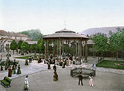 A gazebo-structure with drinking fountains at the base, in a tree-lined square.