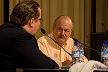 A middle-aged male in a white button-down shirt sits behind a microphone at a convention.