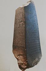 Photographie d'une stèle pyramidale à base rectangulaire, en pierre foncée. Les deux faces visibles sont gravées de nombreuses inscriptions.