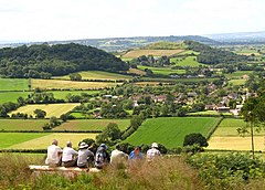 A patchwork of green and yellow fields with the roofs of houses visible centrally. In the background are hills and in the foreground the backs of several seated people.