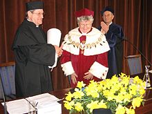 Zdjęcie. Troje profesorów stoi na sali uniwersyteckiej. Norman Davies przyjmuje owacje, ubrany jest w czarną togę akademicką, na głowie czarny biret. W rękach trzyma zwinięty w rulon dyplom doktoratu honoris causa. Obok Katarzyna Chałasińska-Macukow w czerwonej todze z białymi gronostajami. Na jej szyi łańcuch rektorski, na głowie czerwony biret. Kilka metrów za nimi stoi Henryk Samsonowicz, bije brawo. On ma na sobie granatową togę oraz biret.