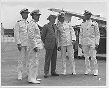 Black and white photograph of five men; two men each in white naval uniform with caps standing to the right and left of a man in a suit with a top hat; a car and an opened hangar are at their right and to the back, a group of men in white naval uniform is at the bottom extreme left