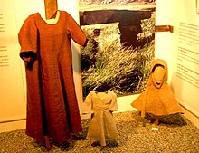 Das Foto zeigt drei in einem Museum ausgestellte Kleidungsstücke, ein langes Kleid, ein Kinderkleid und eine Gugel.