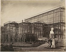 Vue partielle du bâtiment, avec plantations récentes au premier-plan. Un moustachu est appuyé contre le socle d'un buste en pierre. Derrière, une femme en crinoline se promène avec une ombrelle.