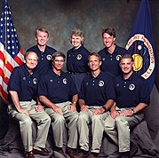 A portrait of six men and one woman, arranged in two rows, four sitting at the front and three standing at the back. They are each wearing tan trousers and a blue polo shirt with a patch and their name on it, and the US and NASA flags are visible in the background.