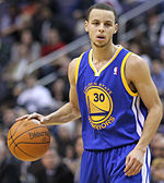 A young man in his early 20s is warming up for a basketball game. He is wearing a red warm-up jersey with "Davidson" written in white across the chest. He is a light-skinned male of mixed race (half white by his mother, half African American by his father)