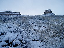 Rocky desert landscape blanketed in snow, shown in near-twilight. Two massifs, several miles in the distance, are snow-covered.