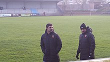 Photo de deux hommes debout devant un terrain de football. Au fond se trouve une tribune de stade.