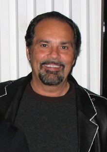 Bruce Sudano wearing a black open jacket with dark top underneath, grinning directly at camera
