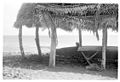 Photo en noir et blanc d'une construction au toit en feuilles de palme sur une plage.