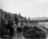 Passenger and goods train under rock ledge beside Tākaka River