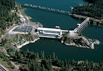 A two-part dam, connected in the middle by an island, blocks a large river downstream of a railroad bridge.