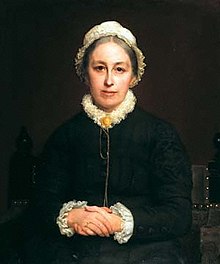 Portrait of woman sitting with hands clasped, wearing a white bonnet and black jacket