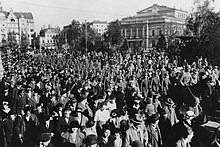 A crowd of civilians are standing in the foreground watching armed soldiers in the middle-ground marching alongside the street. Buildings of downtown Dresden can be seen in the background.