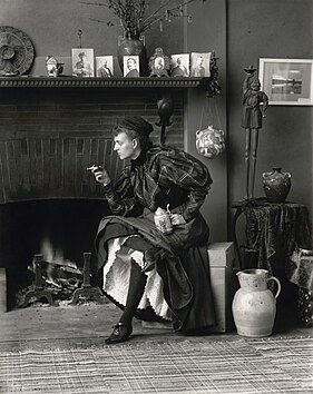 Autoportrait d'une femme assise devant une cheminée, tournée vers la gauche, tenant une cigarette dans une main et une bière dans l'autre.