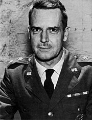 Man with dark hair and moustache in a dress uniform, suit and tie, sitting at a table, with a star indicating his rank, in front of a map on a wall.