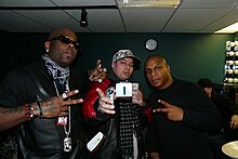 Two African-American men in black attire pose for a photograph, performing hand gestures to the camera