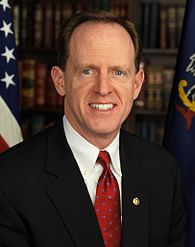 A smiling man with gray hair wearing a black suit jacket and red tie stands in front of the American and Pennsylvanian flags and a bookshelf with several rows of books.