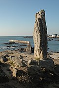 Photographie d’un menhir dressé, un port maritime en arrière plan.