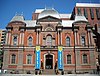 A 3-story, brick and red stone, second empire style building with a slate roof. The building is symmetrical with the entrance center and a blue and yellow banner on either side.