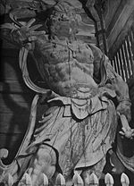 Ungyō. Front view of a scary statue with the right arm raised and carrying a weapon in his lowered left arm. Sculpted breastplate and necklace are visible.