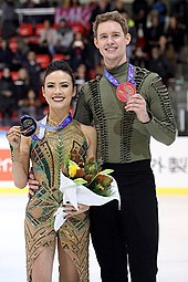 Madison Chock und Evan Bates stehen auf der Eisfläche, Chock links, Bates rechts leicht hinter ihr. Sie tragen ihre Kostüme: Bates ein khakifarbenes Hemd zu einer schwarzen Hose, Chock ein goldenes, teils transparentes Kleid mit grünen und roten Details. Sie zeigen ihre Medaillen vor, die sie an Bändern um den Hals tragen. Chock hält einen Blumenstrauß in der Hand. Im Hintergrund ist das Publikum zu sehen.