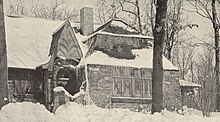 Sepia image of a small snow-covered stone cottage surrounded by tall trees