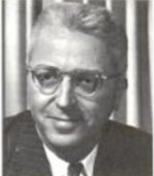 A smiling white man with white hair, wearing glasses and a suit